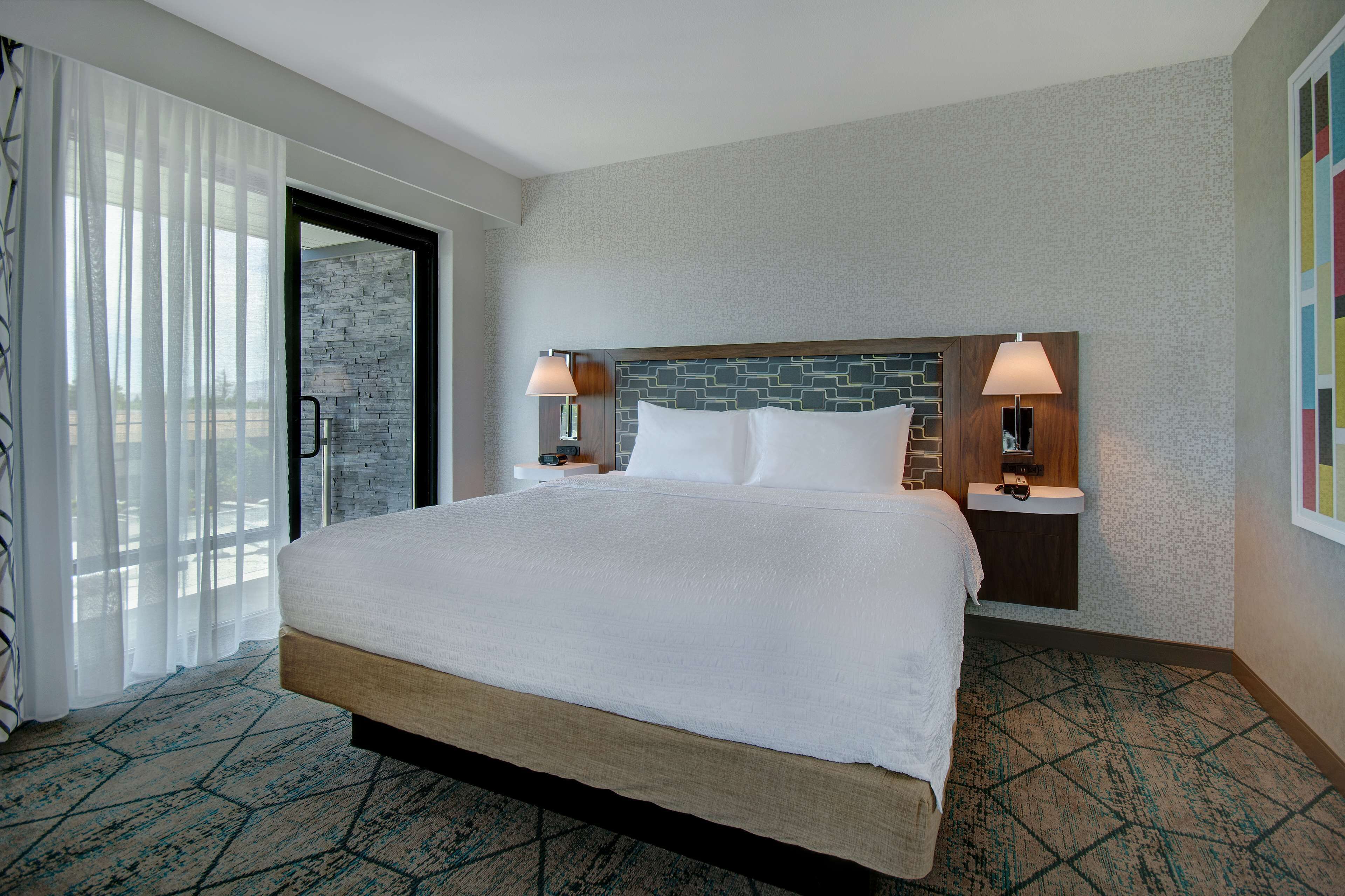A hotel room with a large bed, two bedside tables with lamps, a stone accent wall, and a view through a glass door.