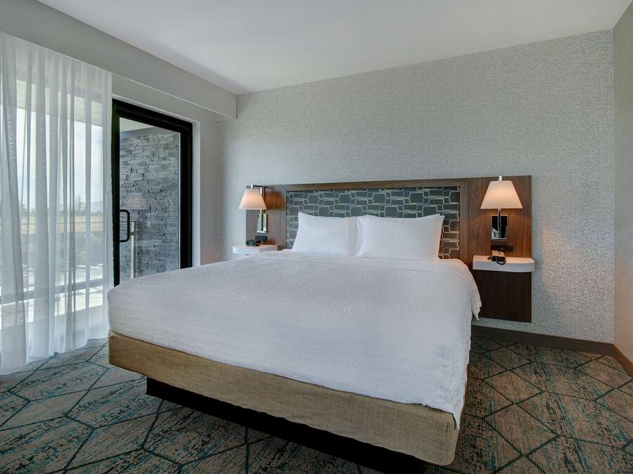 A hotel room with a large bed, two bedside tables with lamps, a stone accent wall, and a view through a glass door.