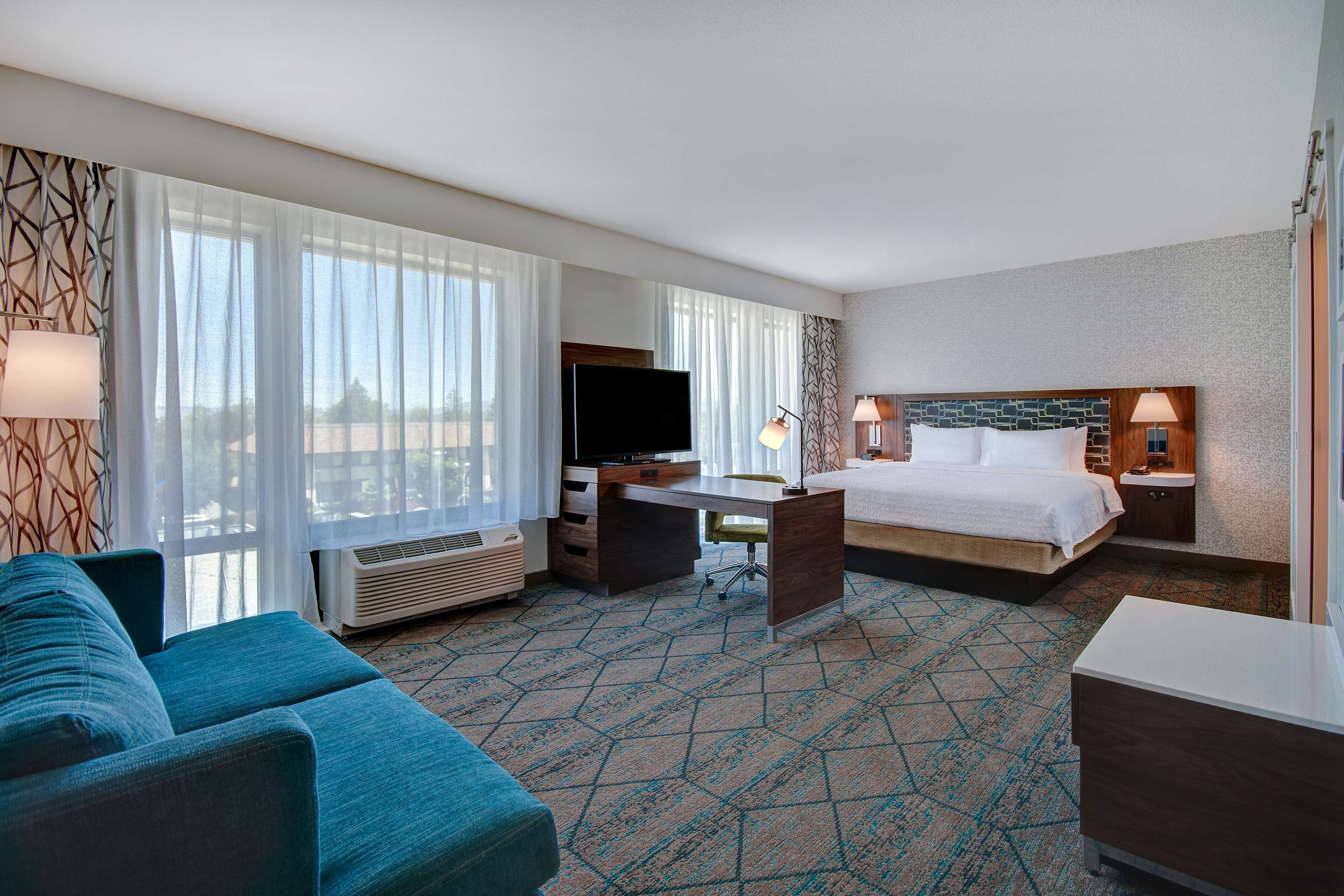 A hotel room with a bed, a desk, a TV, a sofa, and a large window with curtains.