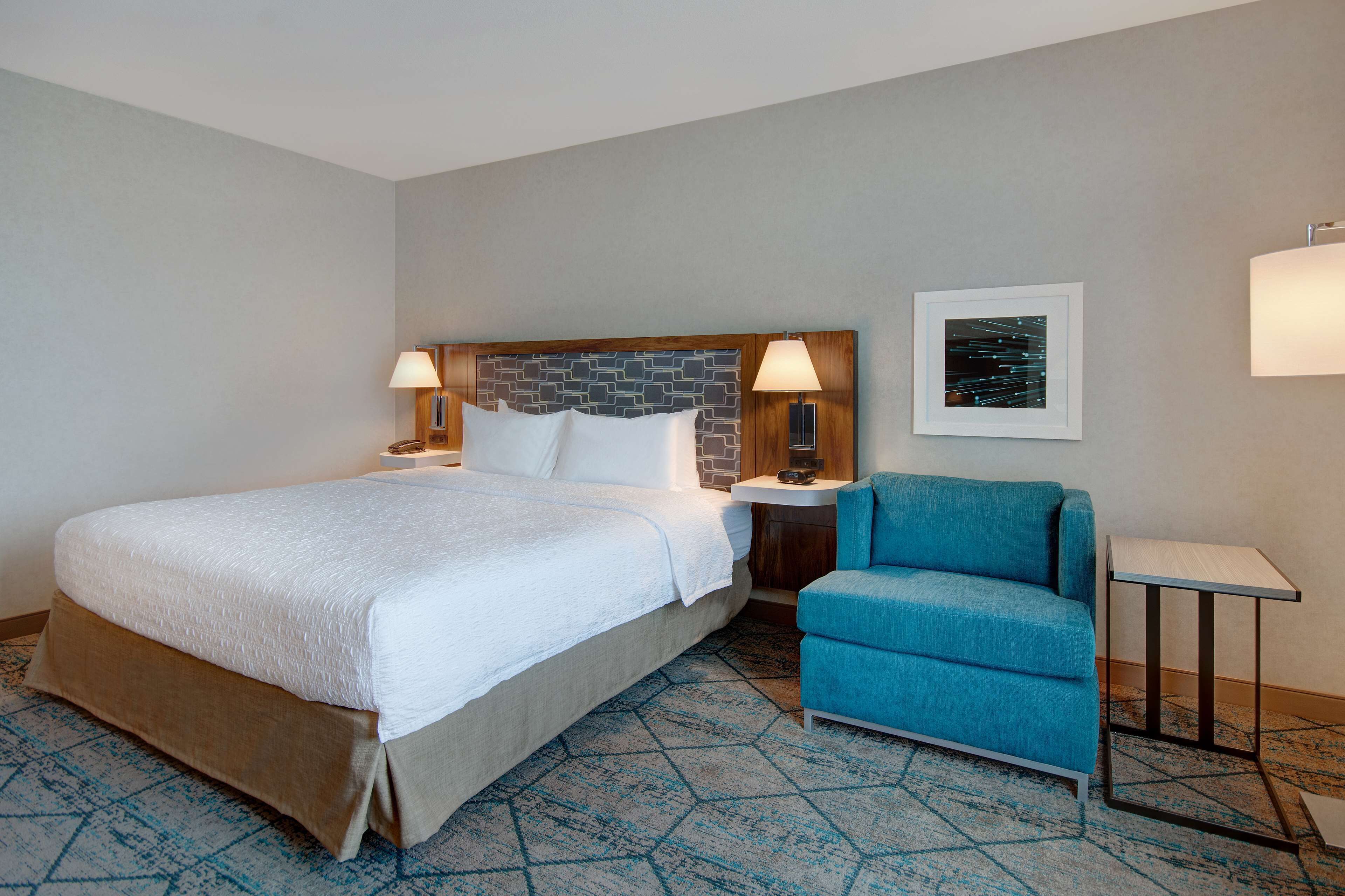 Una habitación de hotel con una cama grande, un sillón azul, una mesita y dos lámparas de noche.