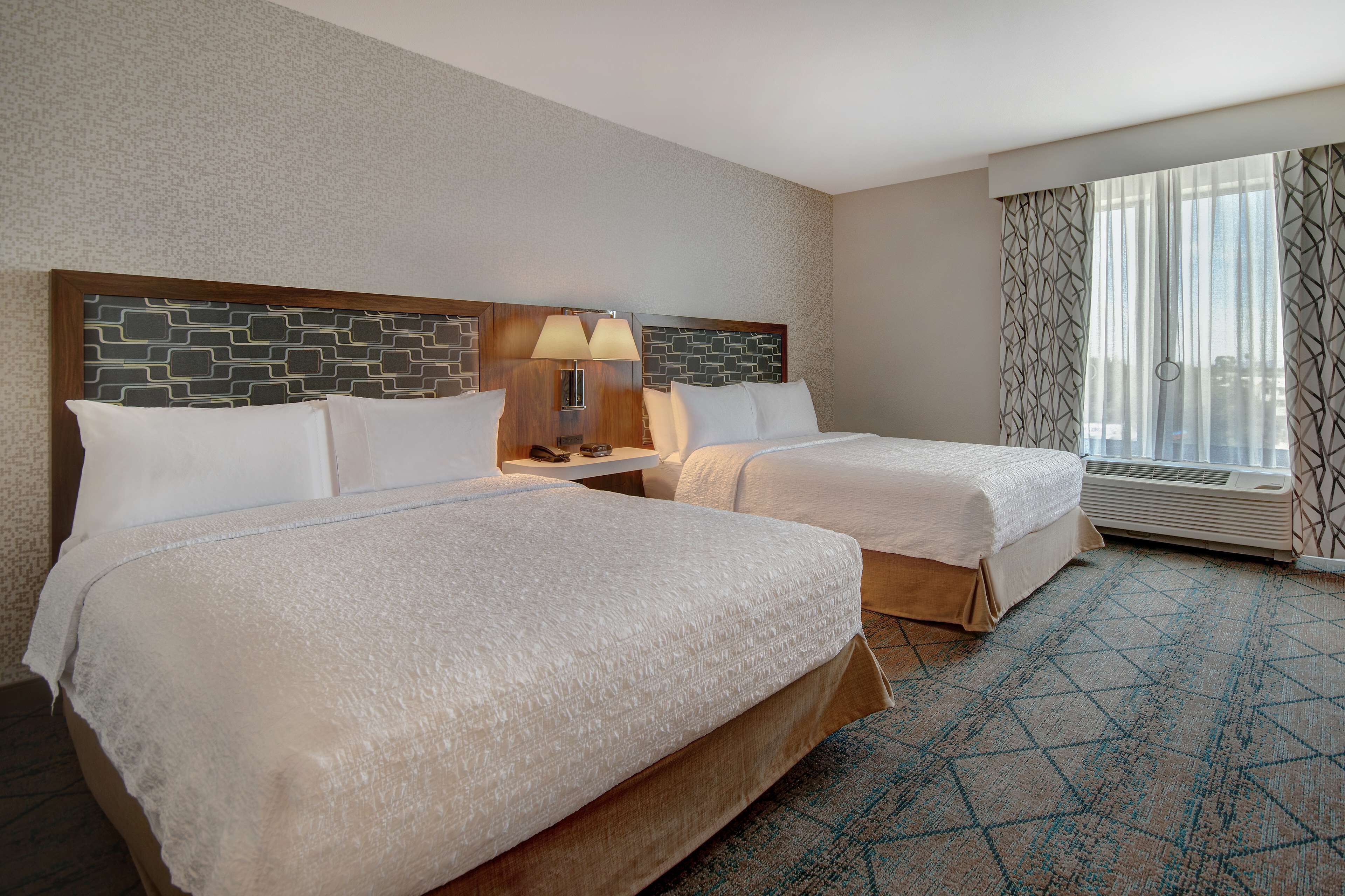 A hotel room with two beds, a nightstand with a lamp, a window with patterned curtains, and a view of the city.