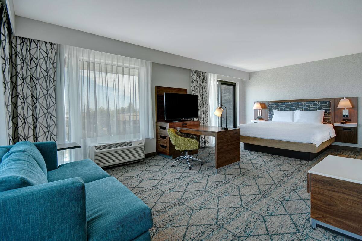A hotel room with a bed, a desk with a TV, a couch, and a view of the city.