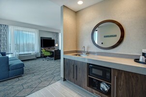 Una habitación de hotel moderna con kitchenette, un espejo grande, televisor de pantalla plana, sofá y vistas al exterior.