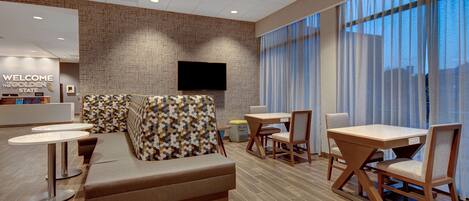 A modern hotel lobby with a reception desk, a sofa, a dining area with tables and chairs, and large windows with curtains.