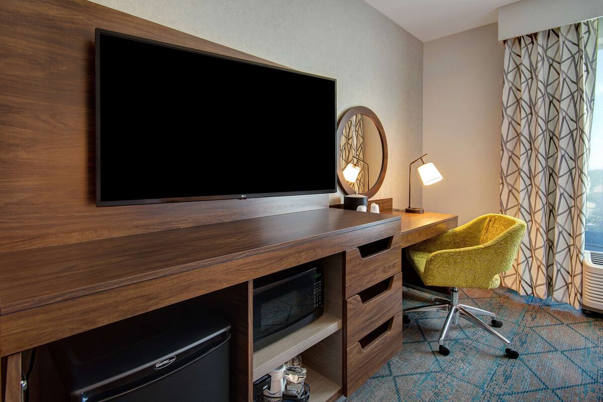 A hotel room with a flat-screen TV mounted on a wooden cabinet, a microwave below it, a desk with a lamp, a yellow chair, and patterned curtains.