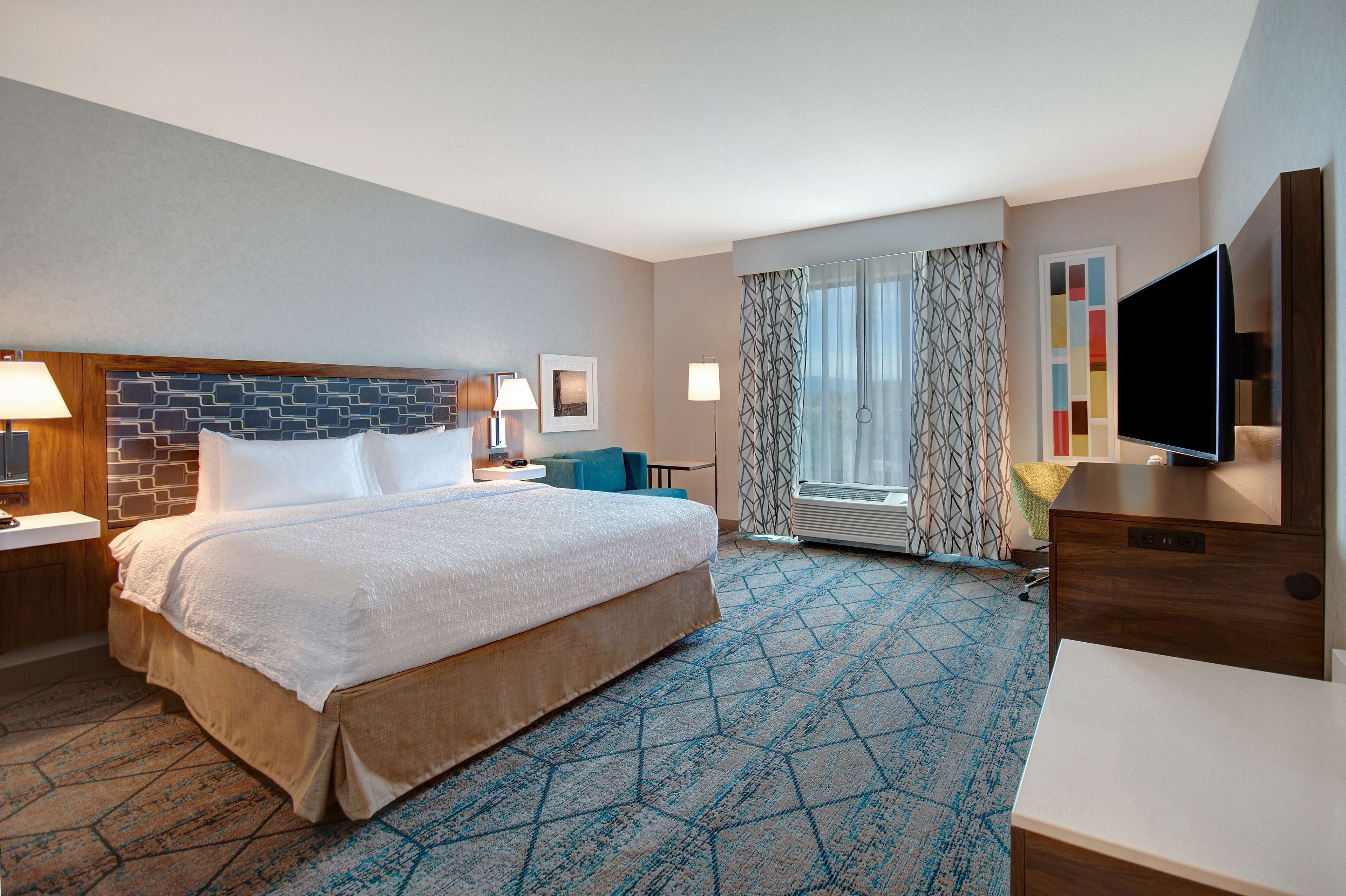 A hotel room with a large bed, a desk with a flat-screen TV, a window with patterned curtains, and a wall-mounted TV.