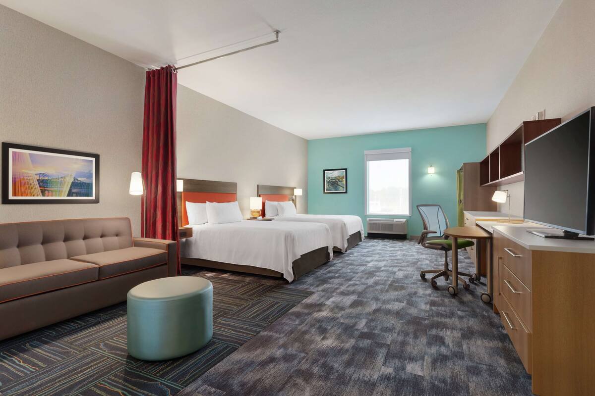 A hotel room with a sofa, two beds, a desk, a TV, and a large window.