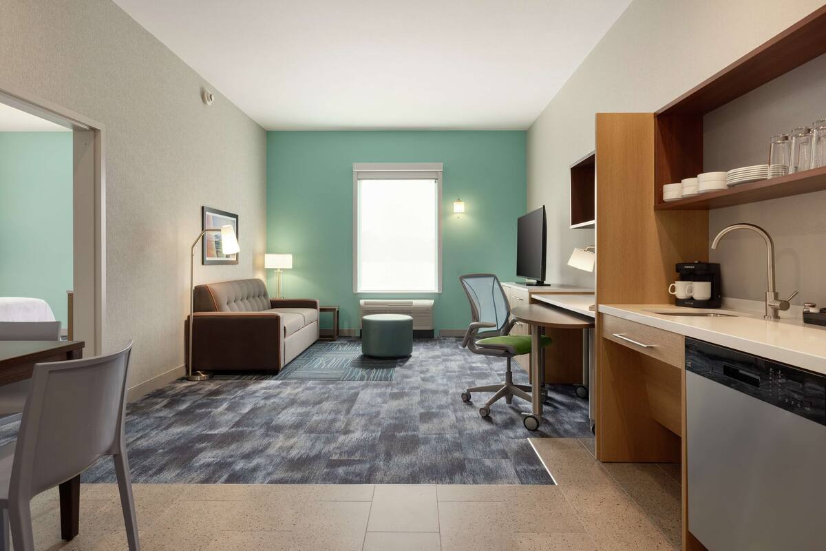 A compact hotel room with a bed, a sofa, a desk with a computer, a chair, a small dining table, and a kitchen area with a dishwasher.