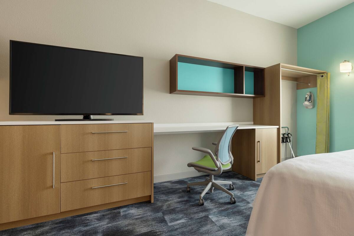 A hotel room with a large flat-screen TV, a desk with a chair, a wardrobe, and a bed.