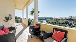 Un balcón con mobiliario de mimbre y cojines rojos, con vistas a un paisaje urbano con palmeras y edificios.