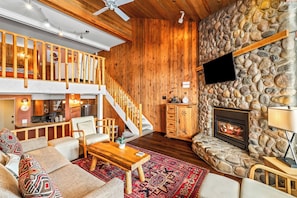 A cozy living room with a stone fireplace, wooden flooring, a staircase, and a TV mounted on the wall.