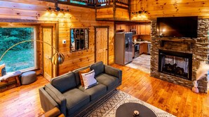 Interior de uma cabana de tronco com sala de estar, cozinha e lareira de pedra.