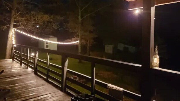 Une terrasse en bois avec une rambarde, une bouteille posée sur un rebord de fenêtre et des guirlandes lumineuses.