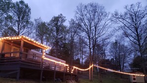 Une terrasse en bois agrémentée de guirlandes lumineuses, un petit abri de jardin et une zone herbeuse.