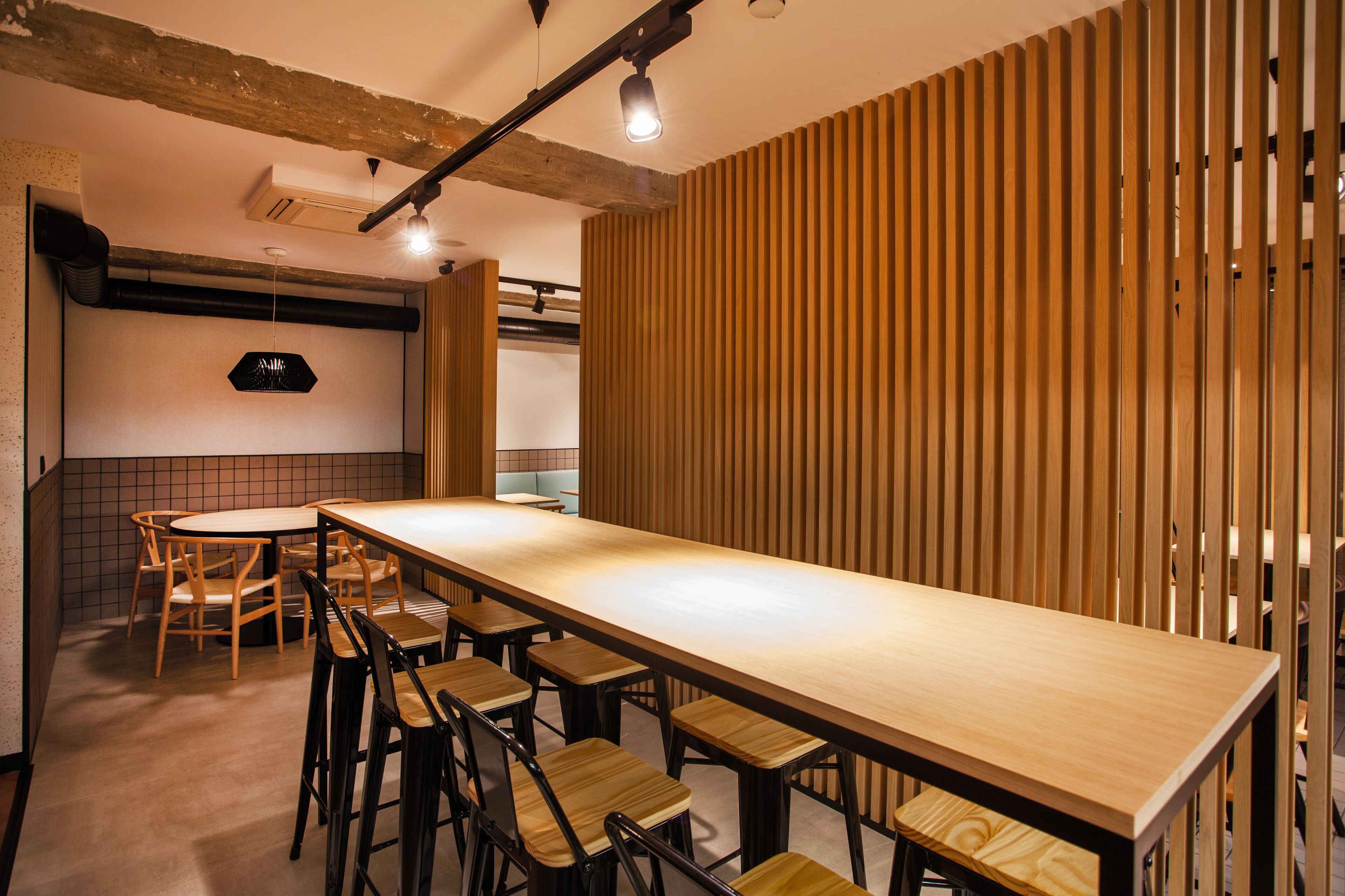 A long wooden table with bar stools, a hanging light fixture, and a tiled wall with a black sign.