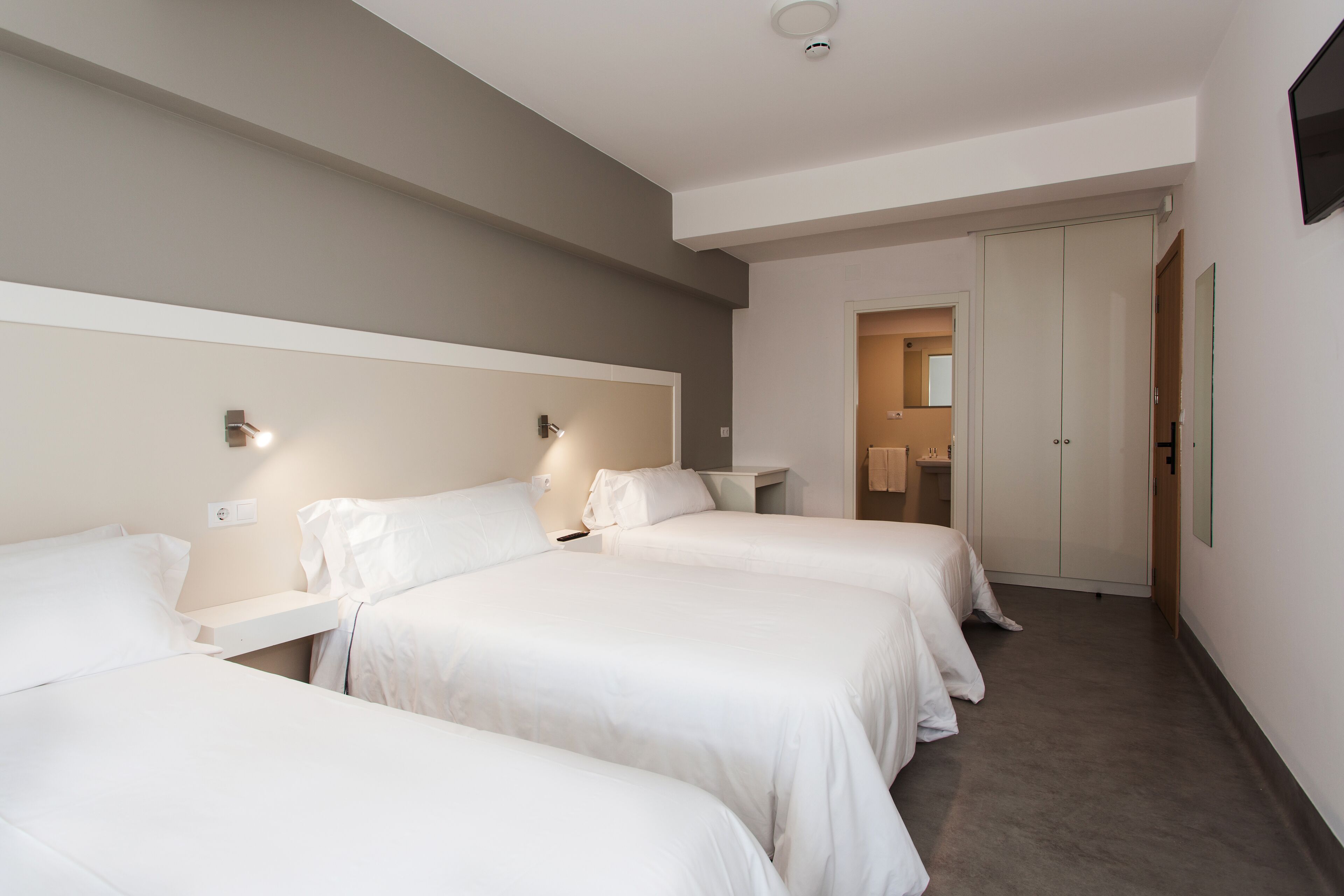 Quarto de hotel com três camas de solteiro, uma mesa de cabeceira, um guarda-roupa e um criado-mudo.