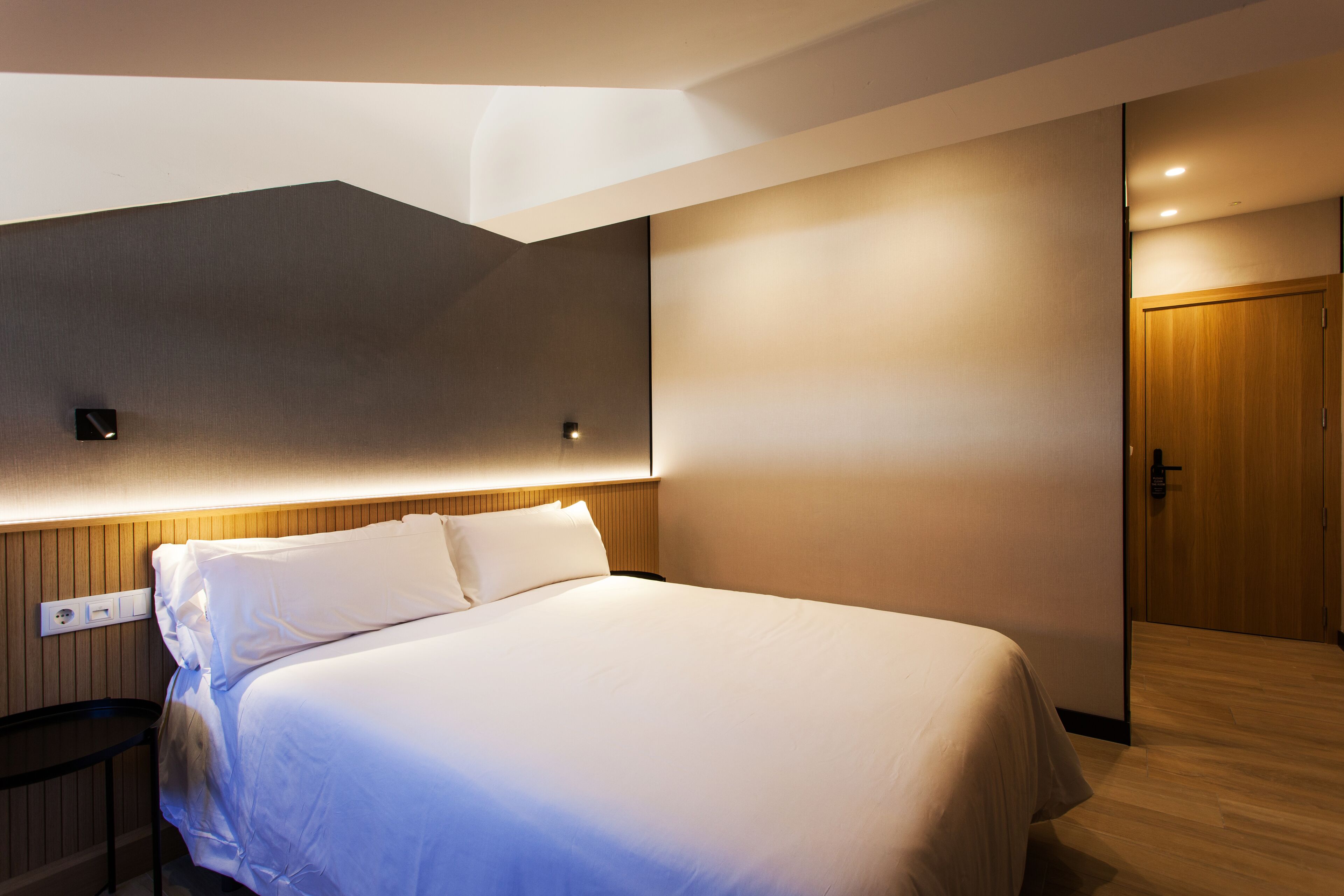 A modern hotel room with a large bed, a wooden headboard, a nightstand, and a wooden door.