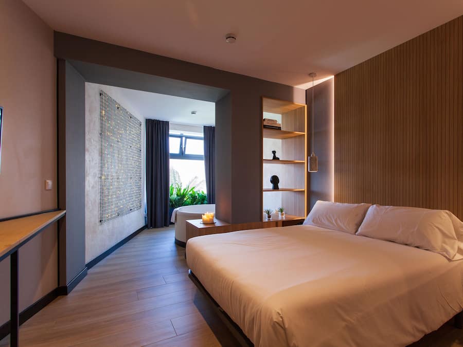 Una habitación de hotel moderna con una cama grande, una mesita de noche, una estantería empotrada y vistas al exterior a través de un ventanal amplio.