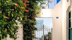 A narrow alleyway with a stone floor, a white wall, and a tree with red flowers. - Room 7 @ Pelican Paros in the Old Town (Paros)
