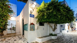 A stone-paved street with a white building featuring a blue sign, a tree, and a lamp post. - Room 7 @ Pelican Paros in the Old Town (Paros)