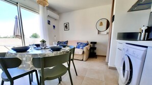 A dining area with a table set for two, a sofa, and a washing machine. - T2 neuf-VUE MER-CLIMATISE-WIFI-PISCINE- 4 personnes-Classé 3*** (Linguizzetta)