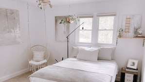 Ein Schlafzimmer mit einem Bett, einem Hängestuhl, einer Stehlampe, einem Fenster und einem Nachttisch mit Bilderrahmen.