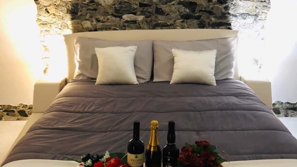 A bed with a stone wall backdrop, a bouquet of red roses, and two bottles of wine. - La Casetta di Angela - Bilocale (Marsala)