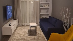 A modern living room with a sofa, a yellow chair, a TV, a white console, and a decorative light fixture. - Flat 66 (Thessaloniki)