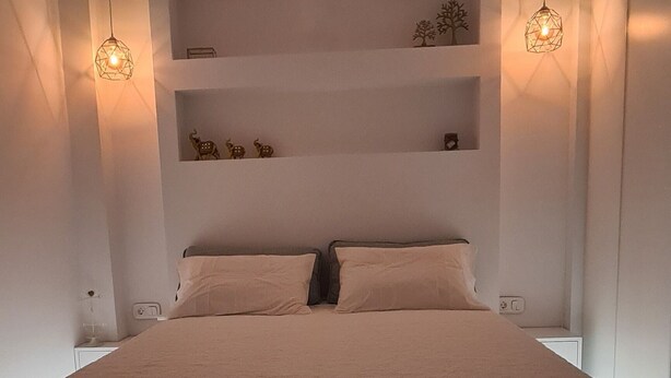 A modern bedroom with a large bed, built-in shelving, pendant lighting, and a recessed ceiling light. - Flat 66 (Thessaloniki)