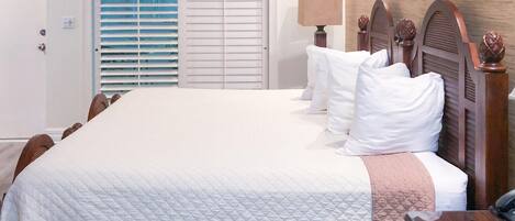 Una cama bien hecha con ropa blanca y cabecera de madera, una mesita de noche con lámpara y una ventana con persianas.