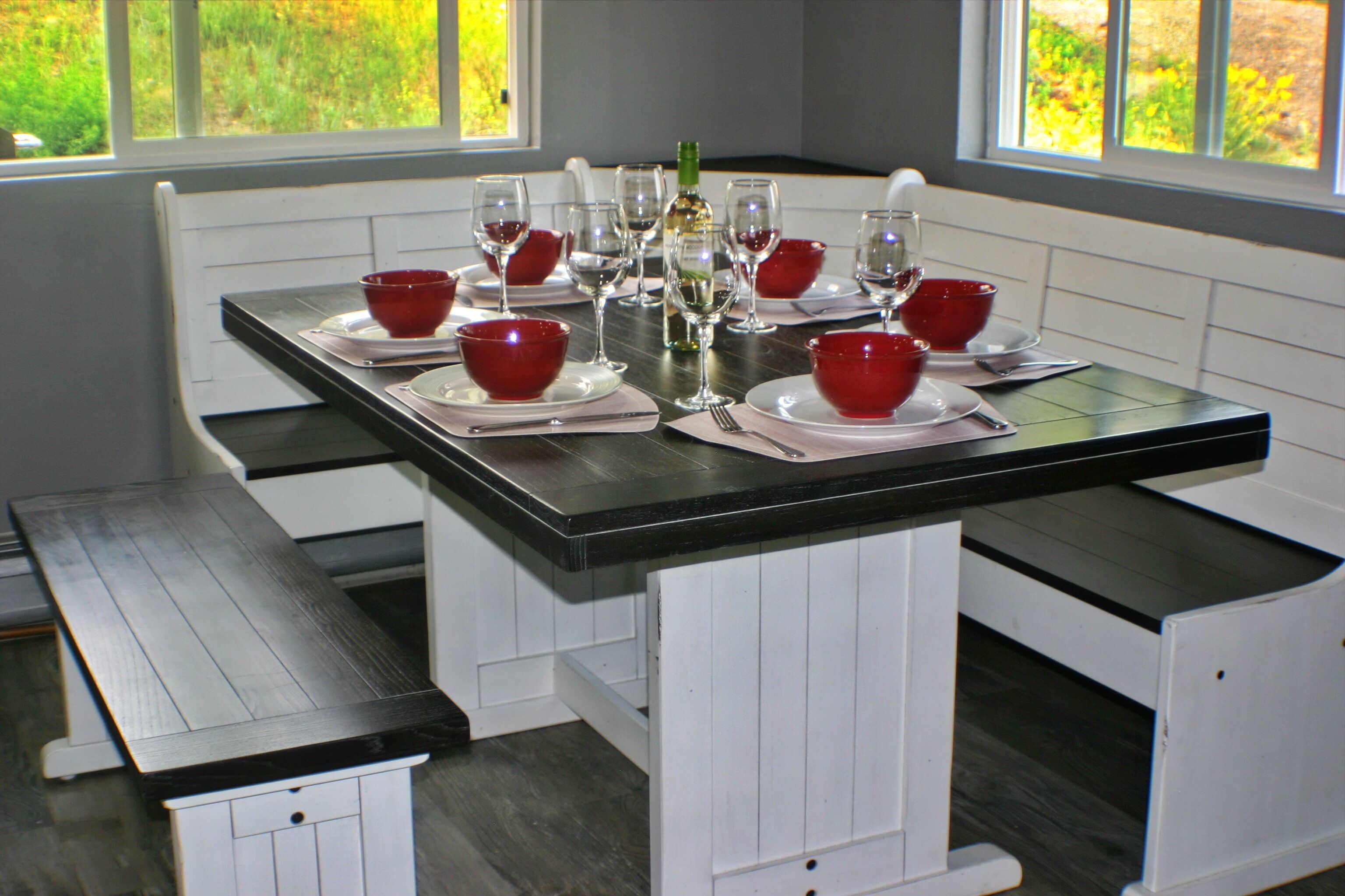 Une table à manger est dressée avec des bols rouges, des verres à vin et une bouteille, le tout entouré de bancs blancs dans une pièce dotée de fenêtres.