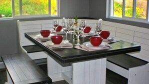 Une table à manger est dressée avec des bols rouges, des verres à vin et une bouteille, le tout entouré de bancs blancs dans une pièce dotée de fenêtres.