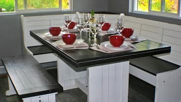 Une table à manger est dressée avec des bols rouges, des verres à vin et une bouteille, le tout entouré de bancs blancs dans une pièce dotée de fenêtres.