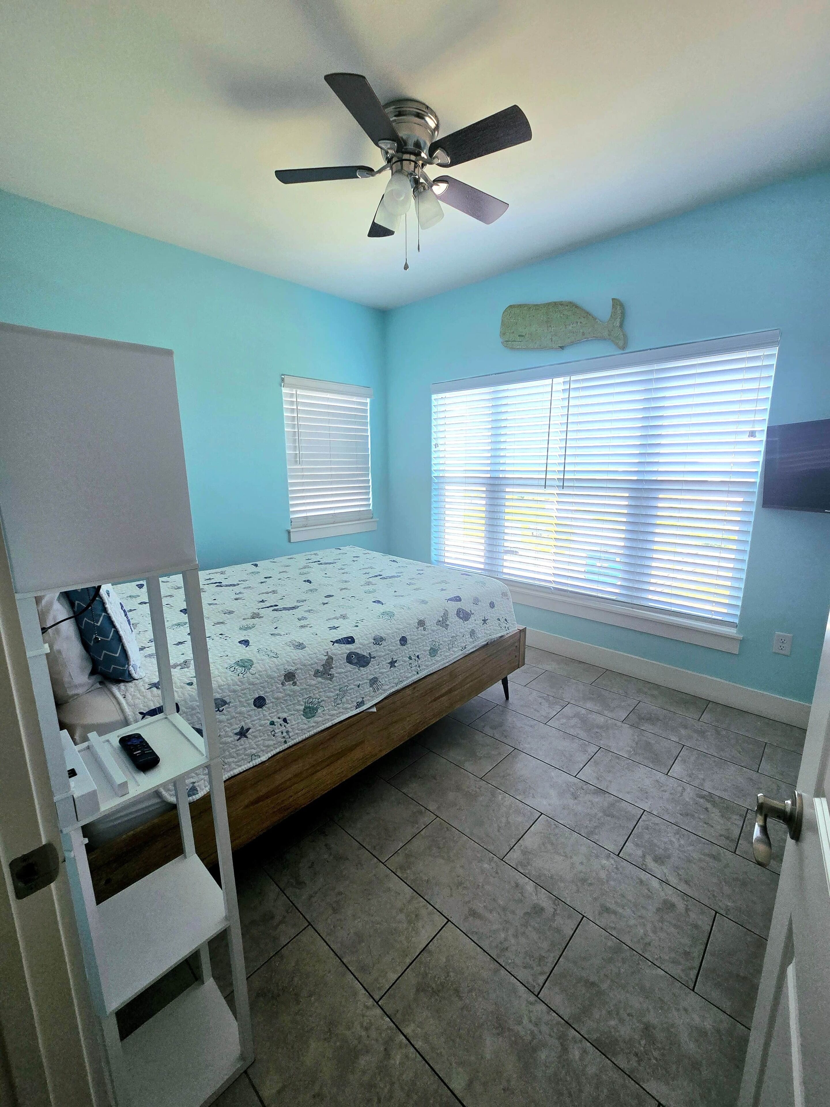 A bedroom with a bed, a ceiling fan, a window with blinds, and a small white shelf with a remote control.