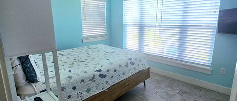 A bedroom with a bed, a ceiling fan, a window with blinds, and a small white shelf with a remote control.