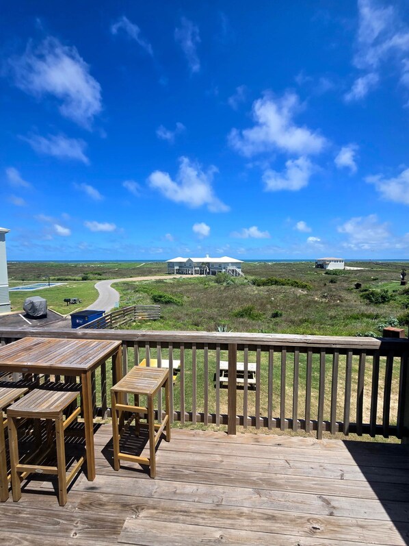 A wooden deck with outdoor furniture, overlooking a grassy landscape and a road leading to a building.