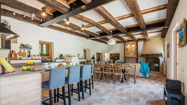 Une cuisine spacieuse avec un haut plafond et des poutres apparentes, un coin repas avec une table et des chaises, et un salon confortable doté d’une cheminée.