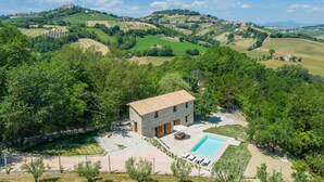 Une maison en pierre avec un toit en tuiles, une piscine et un patio entouré d’arbres.