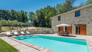 Une piscine entourée de murs en pierre et d’une terrasse en bois, avec des chaises longues et une table à proximité.