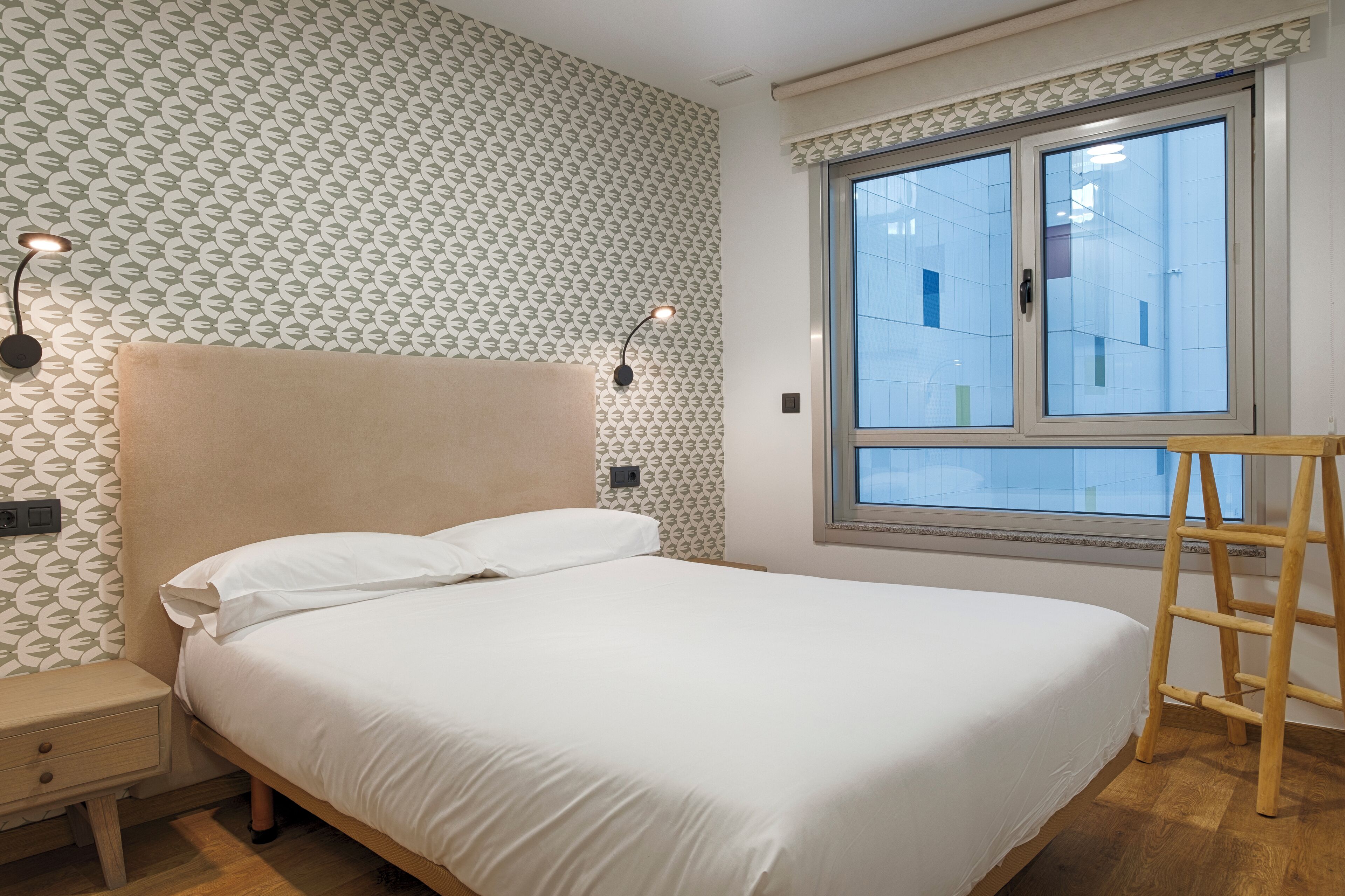 A neatly made bed with white linens, a wooden nightstand with a drawer, two wall-mounted reading lamps, and a window with a view of buildings.