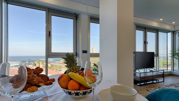 A dining table with a fruit bowl, wine glasses, and pastries, overlooking a scenic view. - TH Apartments by Como en Casa (A Coruña)