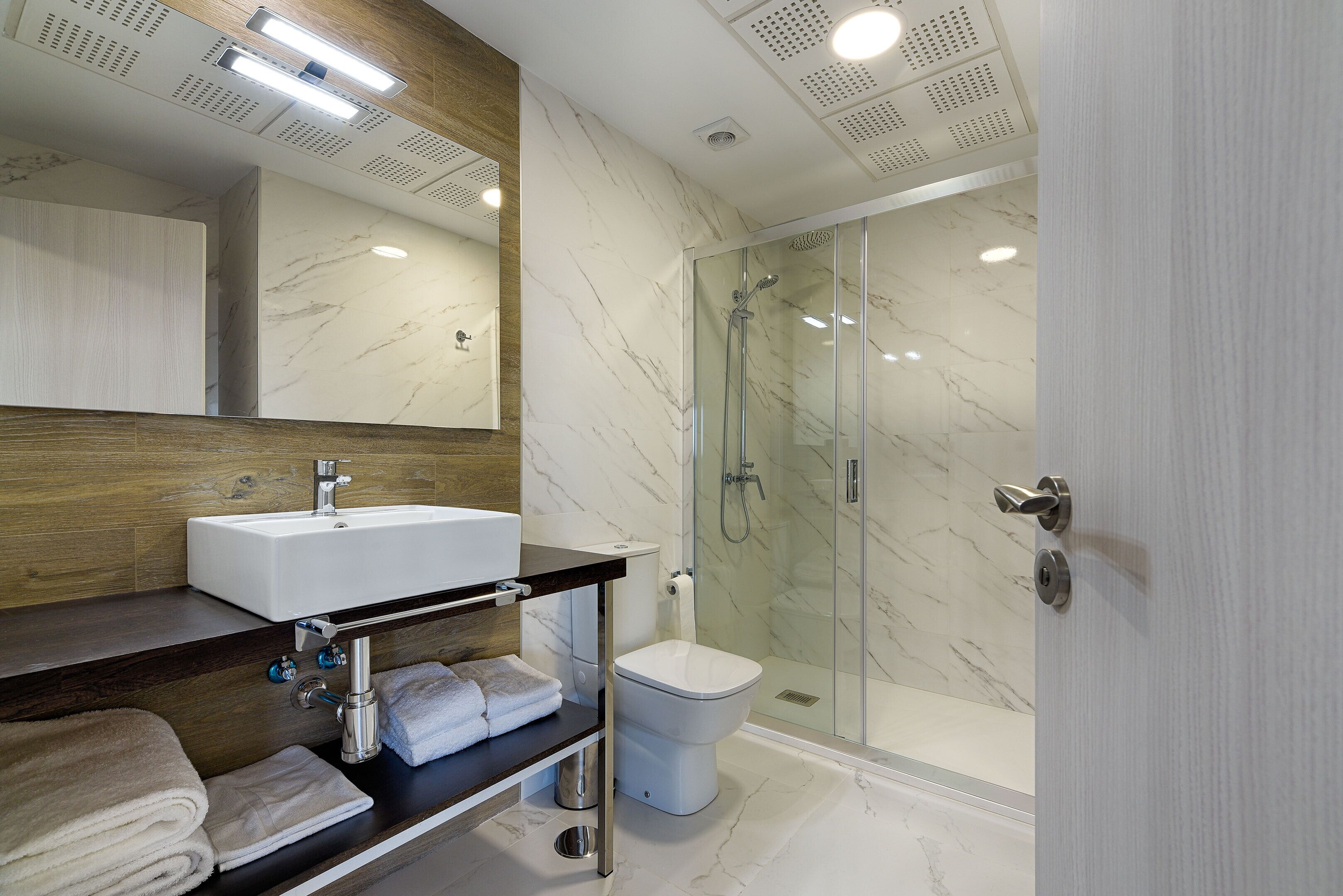 Un baño moderno con ducha de mármol, lavamanos blanco y tocador de madera.