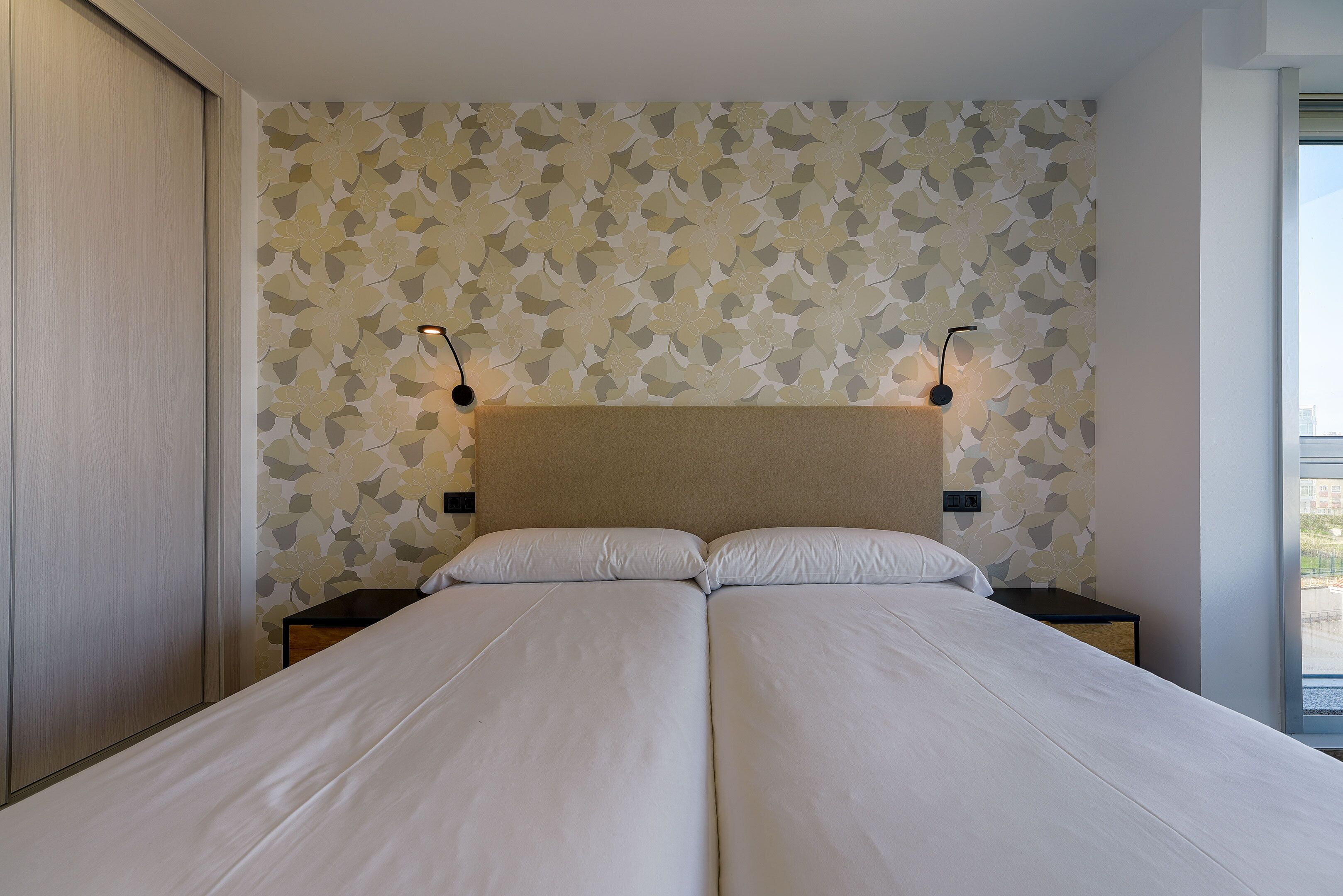 A double bed with a beige headboard, two bedside tables with lamps, and a patterned wallpaper.