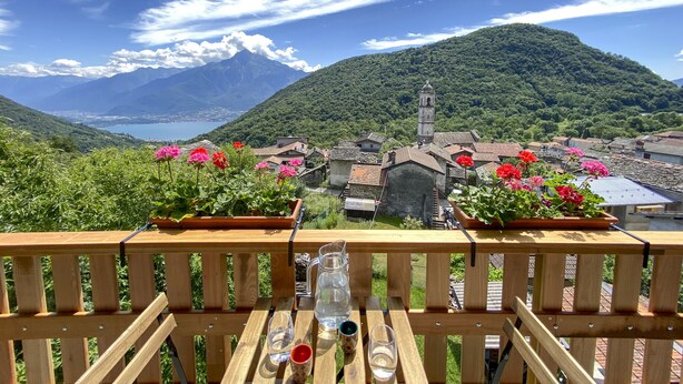 A wooden deck with a table set for two, overlooking a village with traditional buildings and a mountain in the background. - Cosy flat for nature lovers and peace seekers in the mountain village of Livo (Livo)