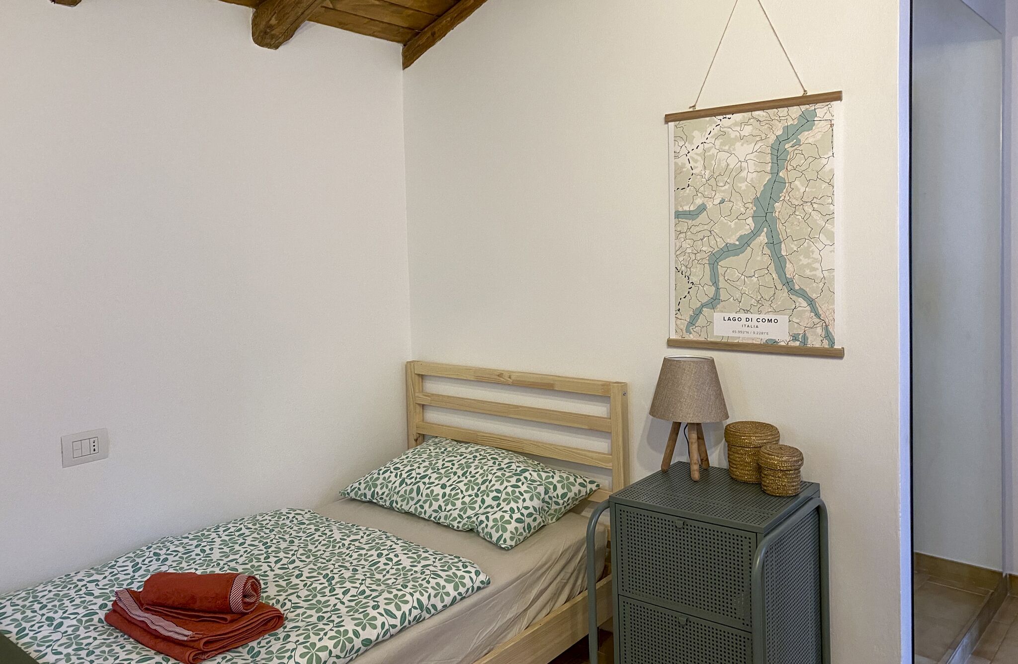 Een enkel bed met een houten hoofdbord, een kaart van Lago Di Como aan de muur, en een tafeltje met een lamp en decoratieve objecten.
