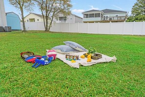 A grassy backyard with a picnic setup including a blanket, pillows, toys, and a small plant.