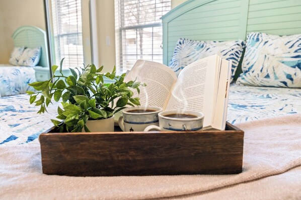 A wooden tray with a plant, two cups of coffee, and an open book on a bed.