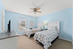 A bedroom with two beds, a ceiling fan, a window with blinds, a desk with a computer, and a wall-mounted TV.