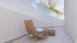 Une chaise en osier avec un coussin, posée sur un sol carrelé, un petit pouf et un mur blanc orné d’un motif géométrique.