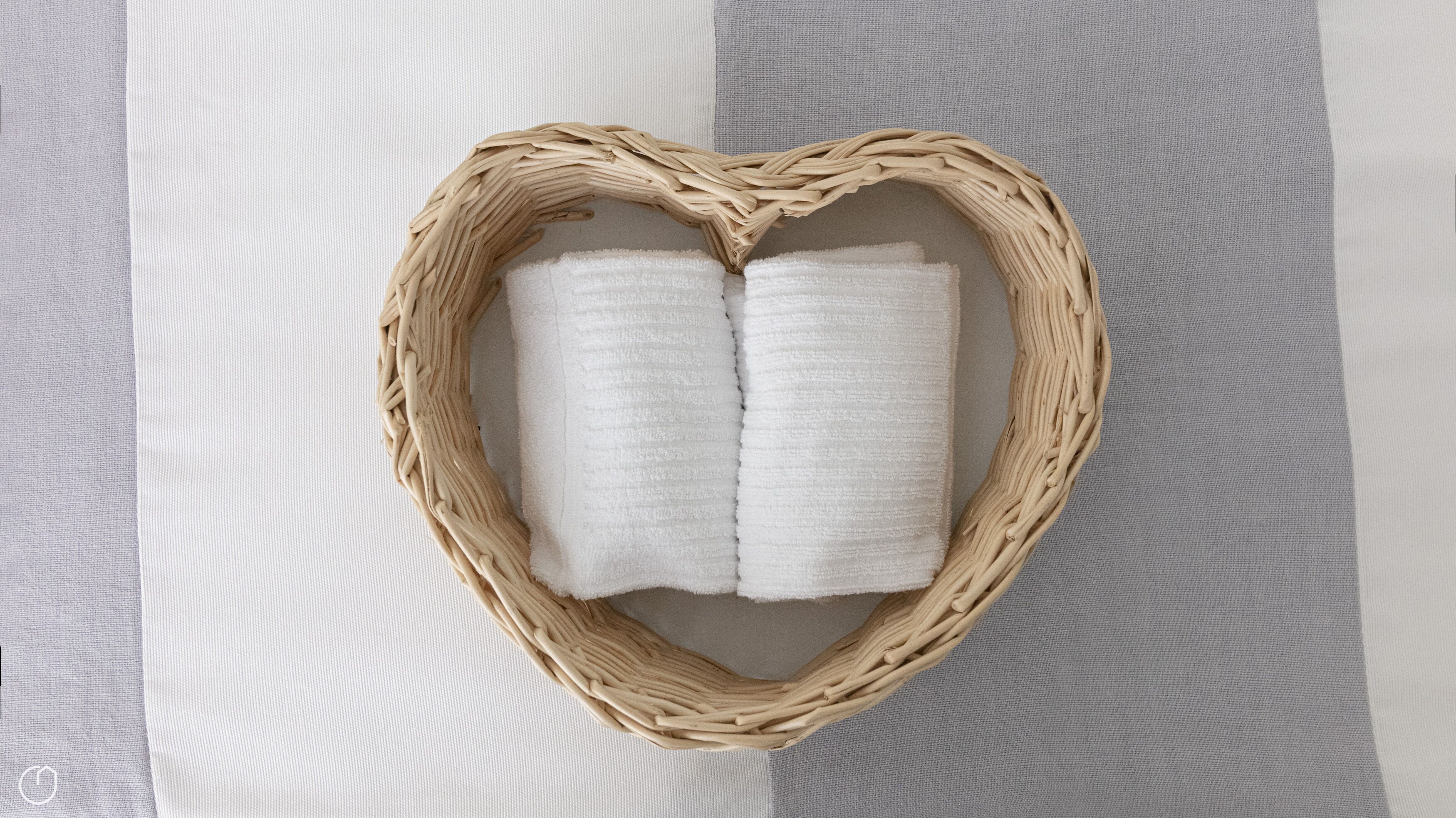 A heart-shaped wicker basket containing two white towels on a textured fabric background.